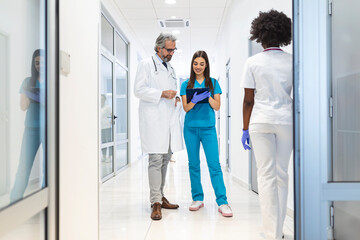 Female Surgeon and Doctor Walk Through Hospital Hallway, They Consult Digital Tablet Computer while Talking about Patient's Health. Modern Bright Hospital with Professional Staff.