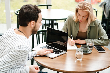 Man and woman using laptop and writing down notes while sitting in cafe