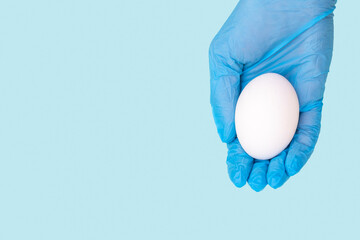 A hand in a protective medical glove holds or gives a white egg on a blue background, copy space, close-up