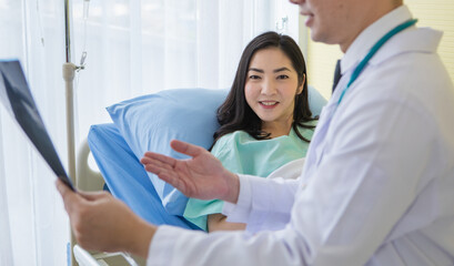 Obraz premium Young Asian medical man in eyeglasses showing chest X ray to smiling ethnic woman lying on sofa in hospital office.