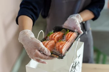 Selbstklebende Fototapeten hand of a waitress filling take away box of sashimi and sushi, alternative activities of catering business in pandemic time, delivery asian food preparation © MandriaPix