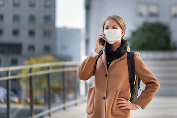 young business woman smiling and talking in a smartphone call, university student wearing facemask, elegant millennial girl in the city