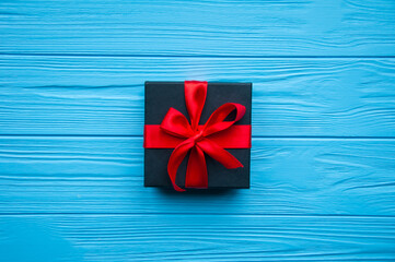 Black gift with red ribbon and bow, isolated on blue background. Present box on wooden board.Gift box with red bow on wood background. Space for text. A gift for Birthday, Wedding, or Valentine's day.
