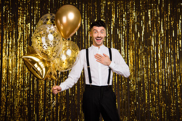 Photo of impressed funky young guy dressed glamour outfit holding many birthday balloons isolated shiny golden background © deagreez