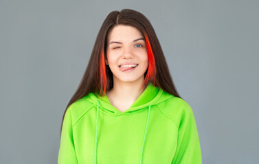 Cheerful young beautiful girl smiling winking showing tongue looking at camera over gray background.