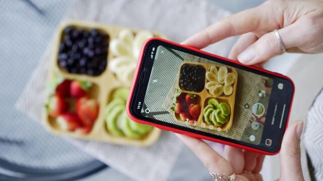 Woman takes picture with smartphone of wooden dish with delicious fresh cut fruits and berries on table in contemporary light kitchen extreme closeup