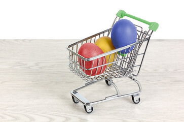 Three colored eggs inside a miniature shopping cart on a white wooden table over a white background