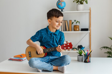 Talented kid playing soprano ukulele sitting on desk. Preschool boy learning guitar at leisure. Concept of early childhood education and music hobby