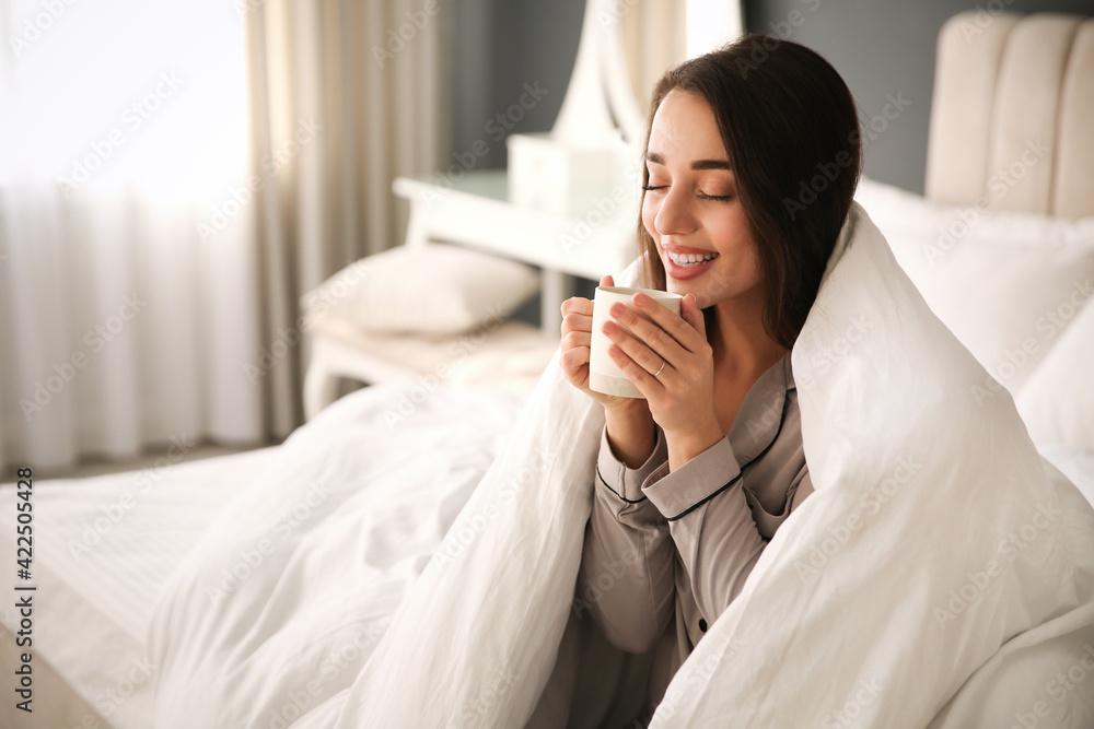 Wall mural Beautiful young woman wrapped with soft blanket holding cup of hot drink on bed at home