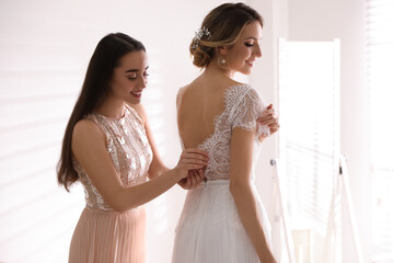 Young woman helping bride to put on wedding dress in room