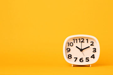 Small white alarm clock, black numbers, set the time placed on a table. Clock on isolated yellow background.