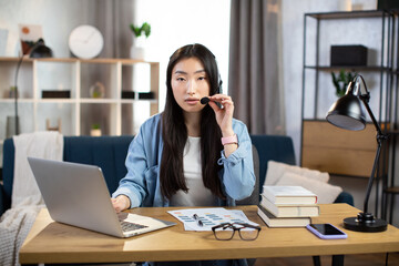 Obraz premium Distance work and learning. Young asian girl student using headset and laptop for video conversation, studying at home. Female freelancer sitting at table and taking notes during video chat.