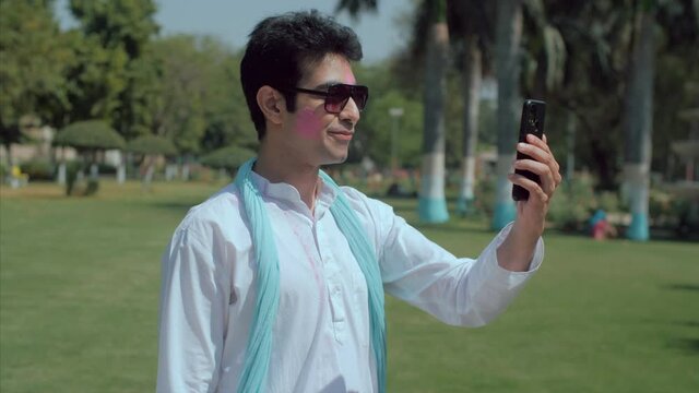 Smiling male adult chatting online during Holi party outdoors - festive mood. Handsome man with Gulal on his face making a video conference call to wish friends on the occasion of Holi