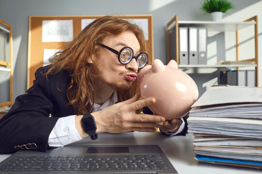 Funny weird looking thrifty man in hilarious round thick lens glasses sitting at office desk with laptop computer and kissing piggy bank. Concept of loving money and saving up