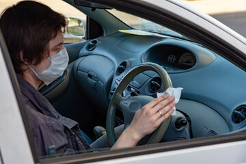 A man in a protective mask wipes the steering wheel with an antibacterial cloth. Infection control concept. Prevent Coronavirus, COVID-19, flu. A man wearing in medical protective mask driving a car.