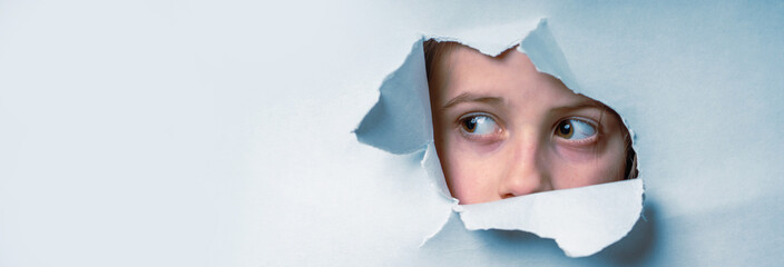 Close up young cute scared child girl  looking through a hole in the paper wall. Horizontal image.