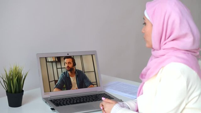 Female islamic student studying online, talking with a male tutor on the laptop screen. Muslim woman wearing hijab connects via video call on the computer with colleague, has virtual meeting