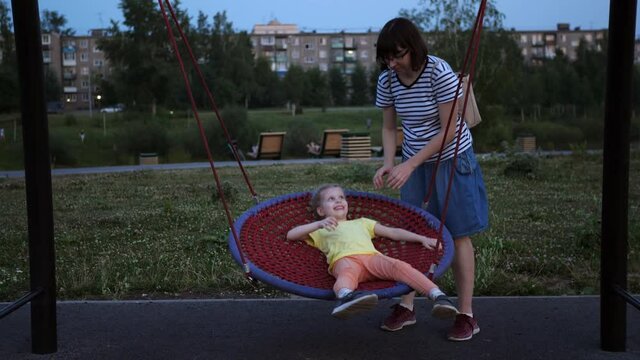 Happy family mom and daughter play on the playground. Young woman tickles child and rocks little girl lying on round net swing in public park at dusk.