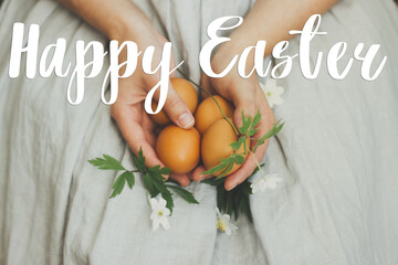 Happy Easter greeting card. Happy Easter text sign handwritten on  woman in rustic linen dress holding natural easter eggs and spring flowers in hands. Beautiful postcard, seasons greetings