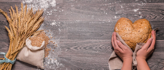 hands hold bread in the shape of a heart, ears of wheat and grain in a bag, dark wooden background, top view, copy space for text