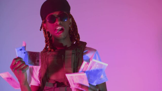 Black man in the studio holds a wad of money and bite the bills with his teeth. A black important man with glasses enjoys wealth. Money flex