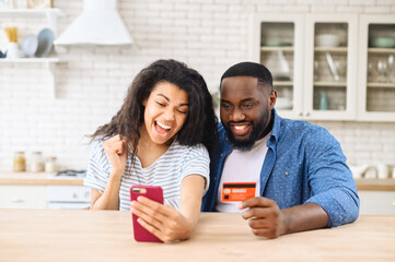 Obraz premium Overjoyed biracial couple shopping together online, excited multiracial woman and man hold smartphone and a credit card, rejoy purchases online, making order, buying something in e-shop