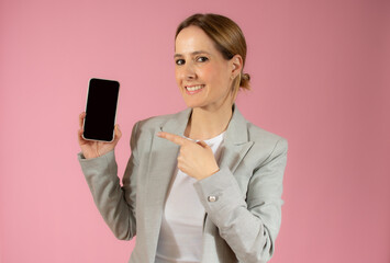 Portrait of a smiling young business woman showing blank screen mobile phone isolated over pink background