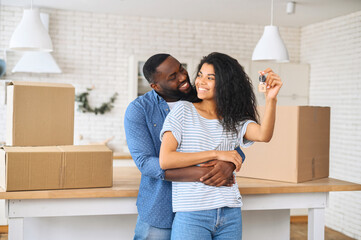Relocation concept. A smiling cheerful multiracial couple in embraces holding keys stating in new...
