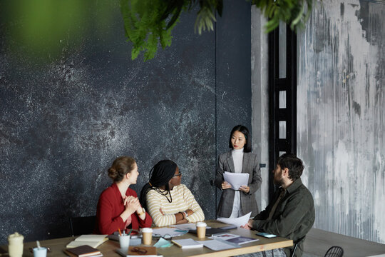 Highh Angle Portrait Of Young Asian Businesswoman Talking To Group Of Colleagues During Meeting At Table In Minimal Grey Office, Copy Space