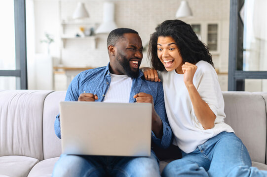 Overjoyed cheerful multiracial couple celebrating good news looking at the laptop screen, young excited woman and man scream happily sitting with computer on the cozy couch at home, won in video game