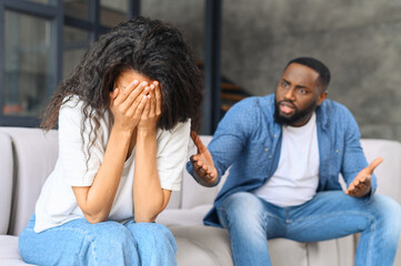 Home abuse concept. Depressed multiracial woman sits on the couch and crying, an irritated and...