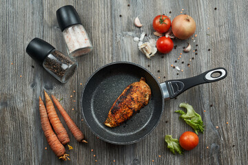 view from above. a black frying pan stands on a gray wooden table in a frying pan there is a well-fried chicken breast. there are vegetables, carrots, onions, tomatoes and mills with salt and pepper 