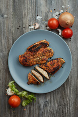 beautiful pieces of fried chicken breast on a gray plate on a wooden gray table. fresh vegetables, tomato, onion, garlic and spices are next to it