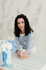 brunette girl with black wavy hair in a blue shirt holding a mug in her hands