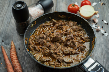 fried chicken liver with onions and carrots in a black pan on a wooden textured table. there are vegetables nearby
