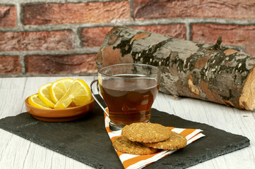 Oatmeal cookies, cup of tea and lemon slices on table over stone plate, wooden log decoration with brick wall background, healthy food concept