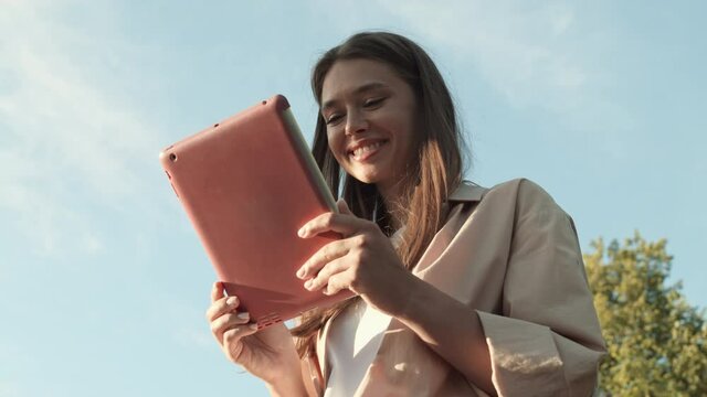 Low angle POV of young delighted Caucasian woman smiling in foreground of green trees and blue sky outdoors, using tablet computer in pink case on sunny day, then looking on camera