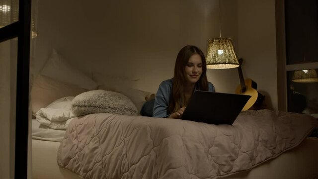 Woman using laptop in bed at home. Joyful woman typing laptop in bedroom. Happy female surfing internet at notebook. Brunette woman in pajamas lying with computer. Attractive lady using laptop in bed