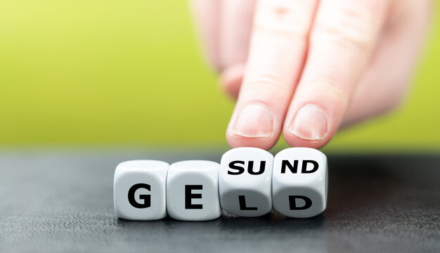 Hand turns dice and changes the German word "Geld" (money) to "Gesund" (health).