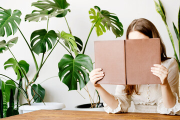 A woman in a light silk dress in a reading room with monstera plants holds a book next to her face © kseniaso