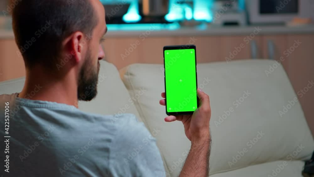 Poster relaxed man looking at smartphone with mock up green screen chroma key display while relaxing on sof