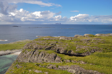 Naklejka premium View from Lovund hills to the rocky coastline and Lovund archipelago in the Norwegian sea on sunny summer day. White clouds over the blue sea. Silhouette of Træna island on the horizon