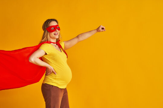 attractive smiling pregnant woman in superhero costume, wearing red mask and cape, stands with her arm outstretched on a yellow background. concept superpowers of a girl, feminism, desire to win
