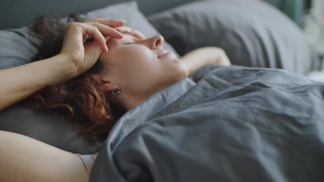 Young woman with curly hair lying on bed, waking up, yawning and rubbing her eyes in the morning at home