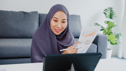 Asia muslim lady wear headscarf casual use tablet talk to colleagues about sale report in conference video call while working from home at living room. Social distancing, quarantine for corona virus.