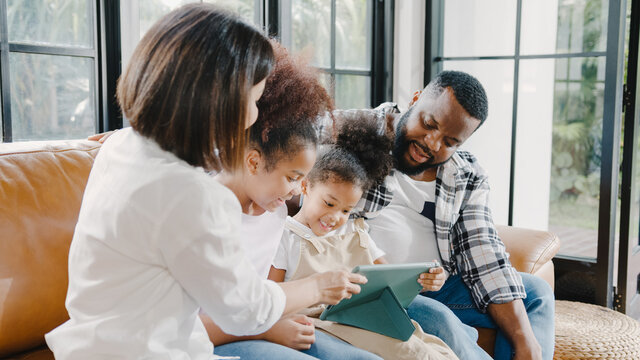 Young African American family and daughter happy using tablet at home. Mom, Dad, and child relax with little girl watching movie on sofa in living room. Funny parent and lovely child are having fun.