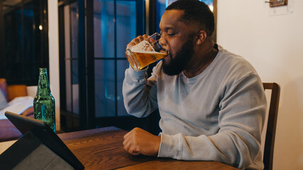 Middle-aged African American male drinking beer having fun happy night party event online celebration via video call in living room at home. Social distancing, quarantine for coronavirus prevention.