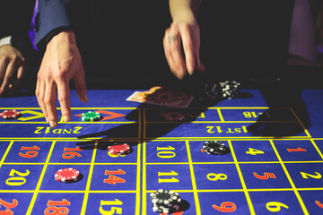 A close-up vibrant image of multicolored casino table with roulette in motion, with casino chips. the hand of croupier, money and a group of gambling rich wealthy people playing in the background