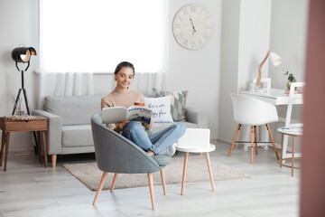 Beautiful young woman reading magazine while sitting in armchair at home