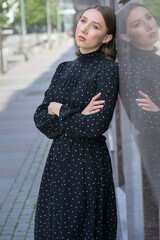 A girl in a black dress with her reflection stands against the wall of a building